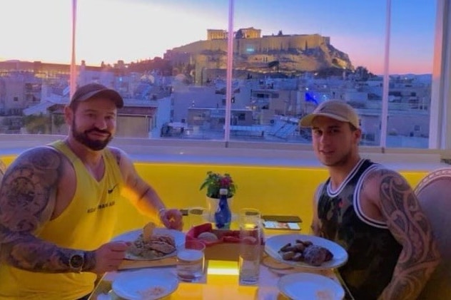 Two men having dinner in a restaurant with the Parthenon through a window in the background.