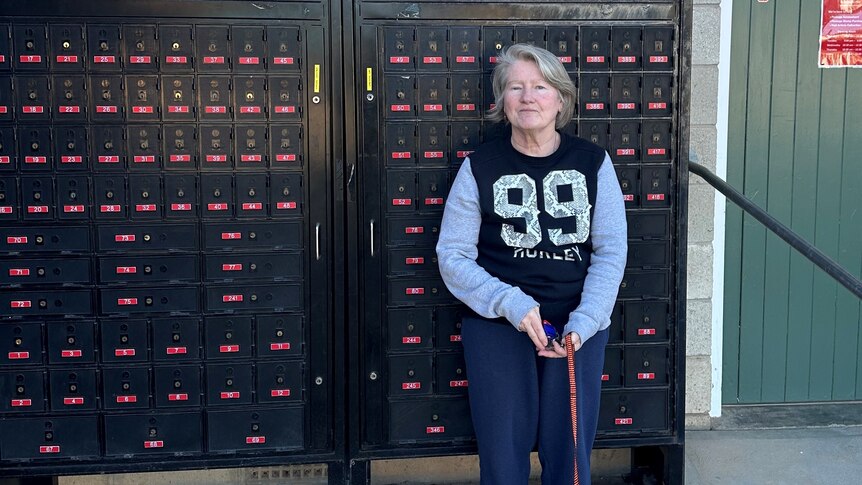 grey haired woman with walking stick standing infront of post office boxes in a wall