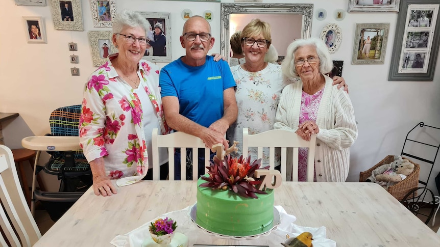 Three older women and an older man stand side by side behind a green cake that says 70 on it.