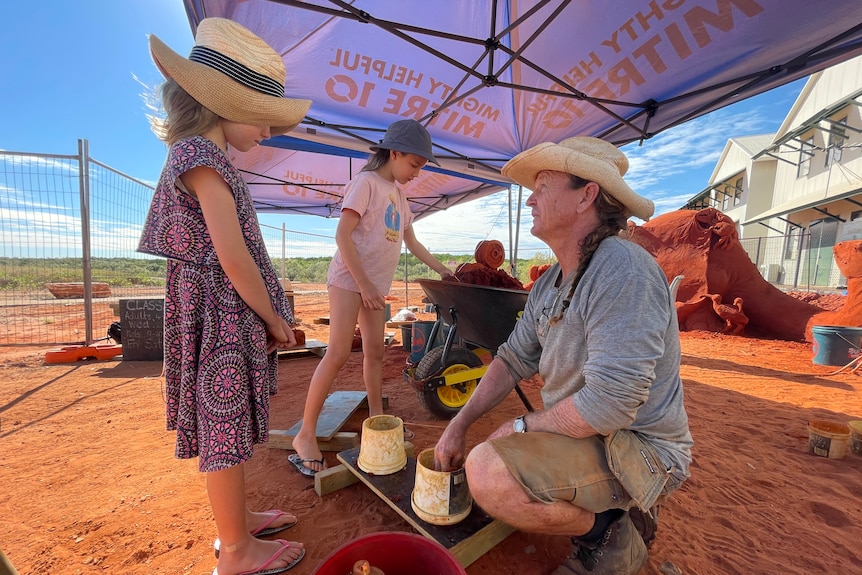 man squatting at a low ledge, demonstrating sculpting to two children standing on red pindan sand, all covered by a sun shade 