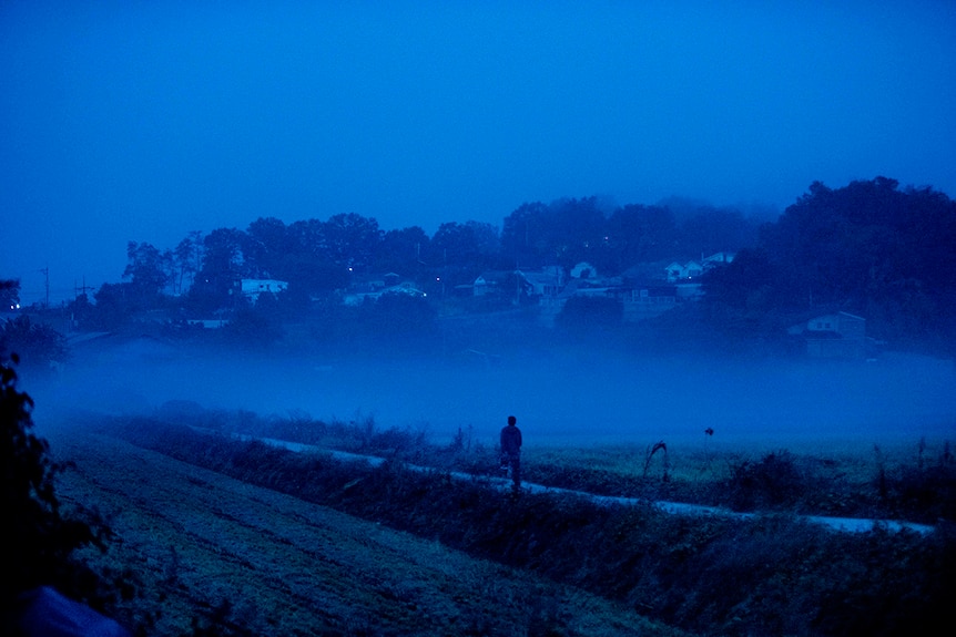 Colour still of Yoo Ah-in jogging in dark blue tinted at night time in rural Paju in 2018 film Burning.