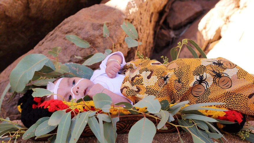 Newborn baby dressed in indigenous colours and fabrics lies with a hallow of gum leaves around her.