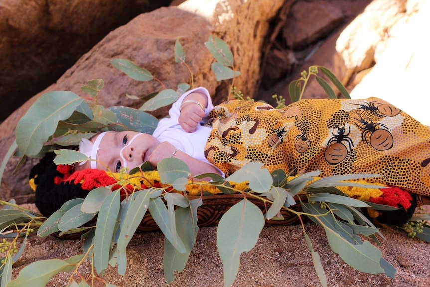 Newborn baby dressed in indigenous colours and fabrics lies with a hallow of gum leaves around her.