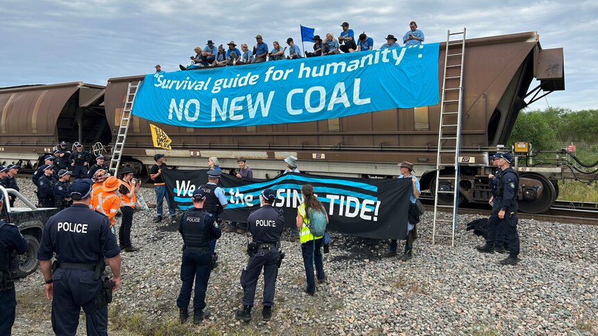 Police stand next to a coal train which has protesters up the top and a large banner "No new coal" draped from the side.