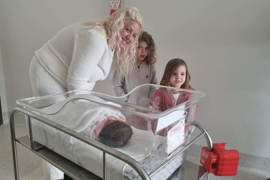 A woman and two girls stand near a crib with a baby