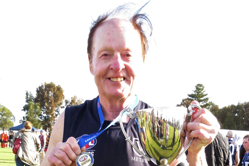 A man in a blue and white Big V footy jumper holds a medal and trophy