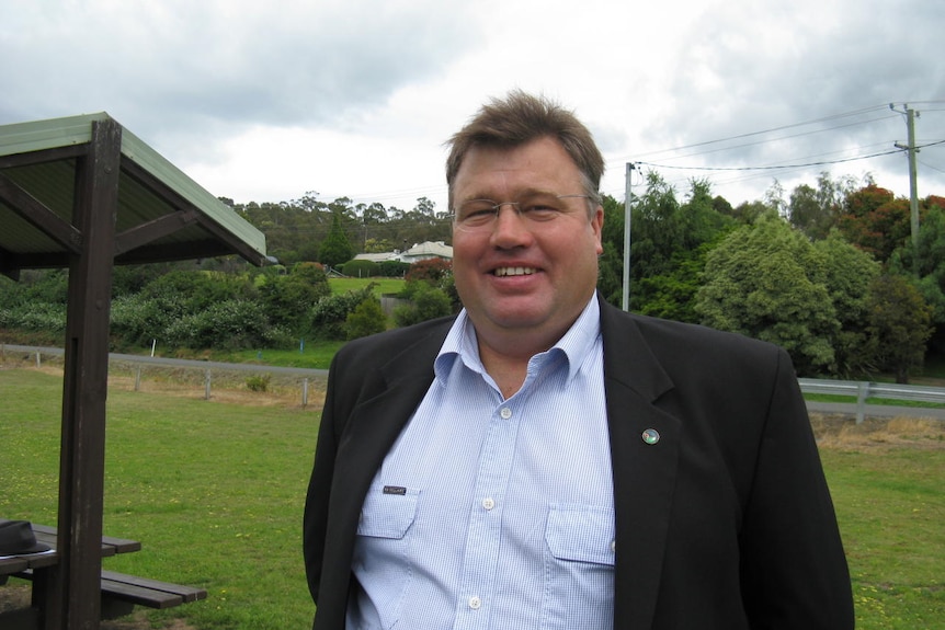 A large man in a white shirt and suit coat stands in a rural setting.