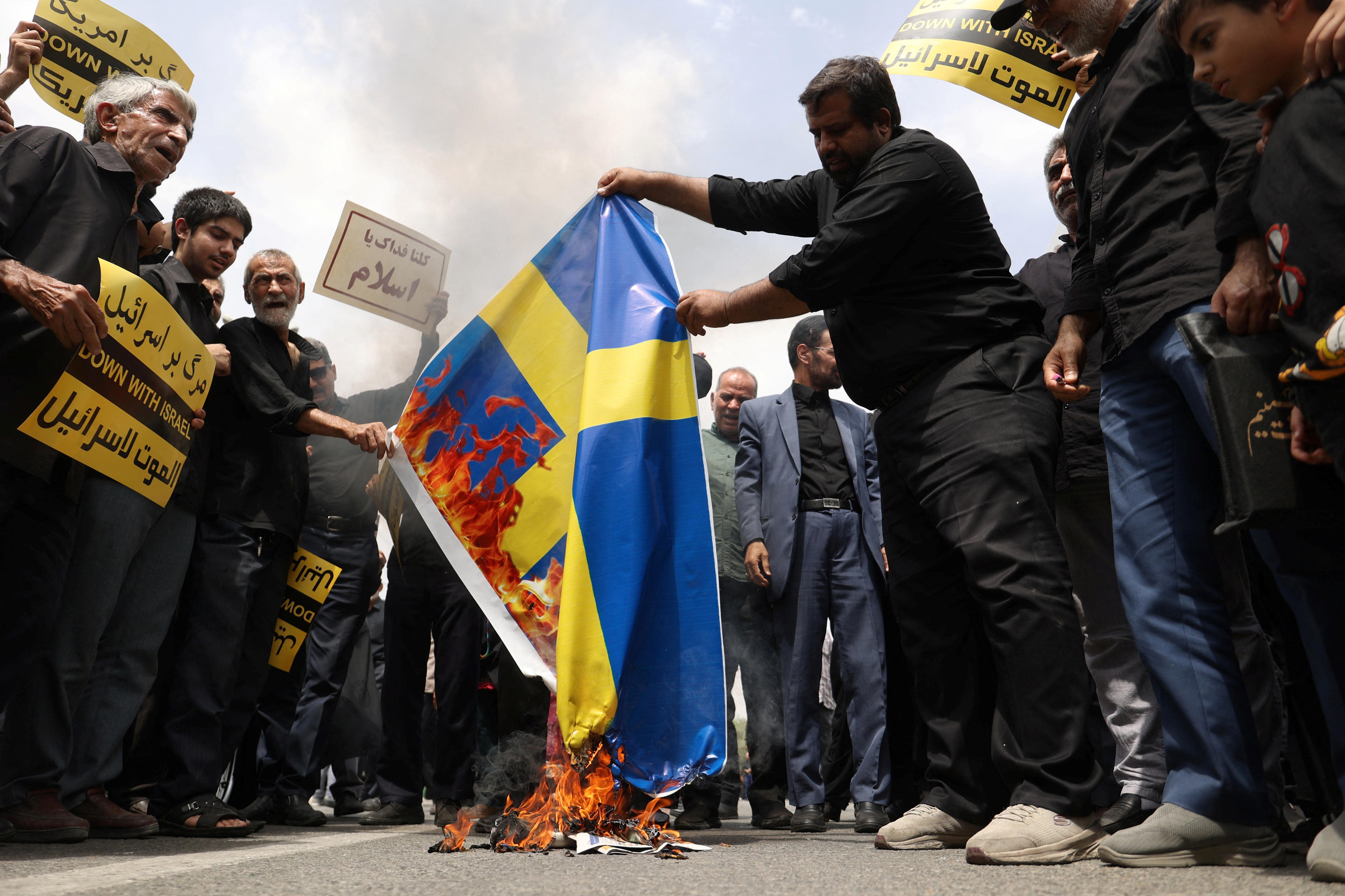 A group of Iranian men stand on a roadway burning a Swedish flag.
