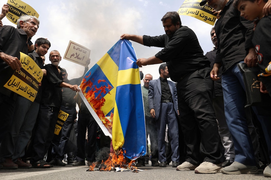 Un gruppo di uomini iraniani in piedi su una strada che brucia una bandiera svedese.