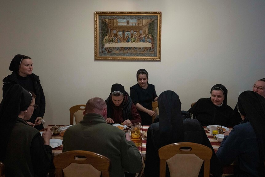 Nuns eating dinner around a table. A framed image of The Last Supper on the wall behind them.