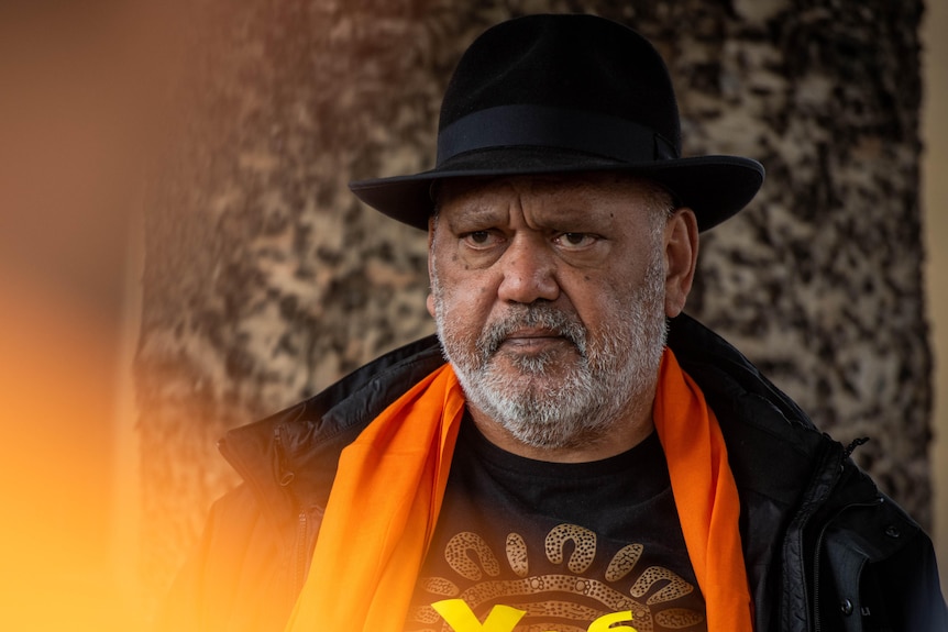 An Indigenous man, white facial hair, with a black hat, wearing a YES shirt, looks to the media