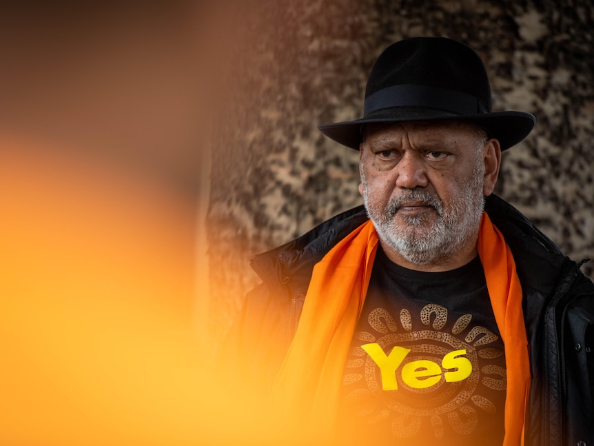 An Indigenous man, white facial hair, with a black hat, wearing a YES shirt, looks to the media