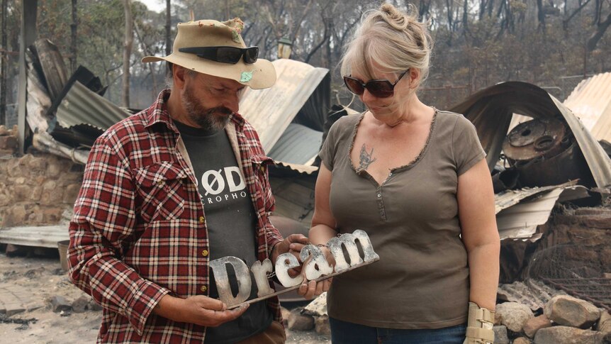 A couple stands in front of a destroyed home, holding a sign that says dream