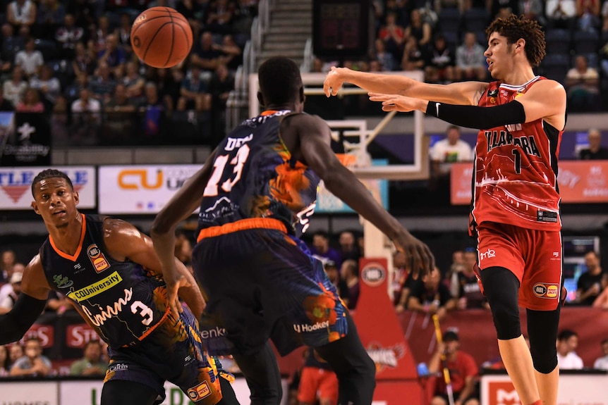 An NBL basketballer passes the ball to a teammate while airborne, as defenders watch him.