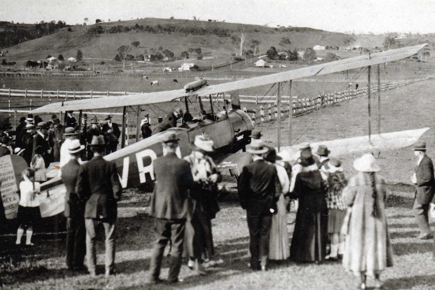 Black and white photo taken in the 1920s of a bi plane in a field surrounded by a crowd of people.