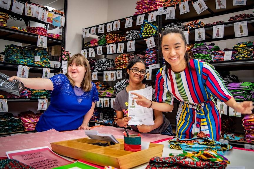 Sophie Grivas and Amrita Ramjas and Stephanie Trinh-Tran laugh and reach their arms out while holding fashion products.