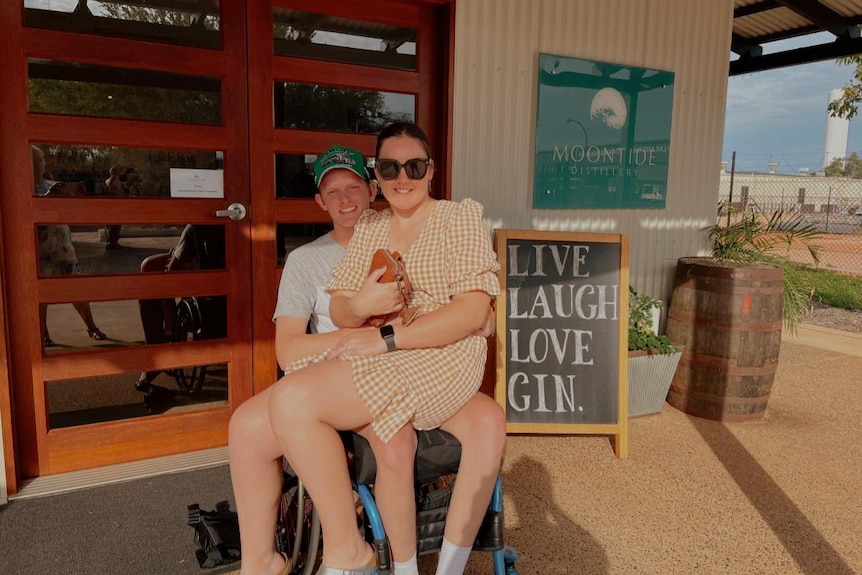 A young woman perches on a man's lap as he sits in a wheelchair.  Both are smiling.