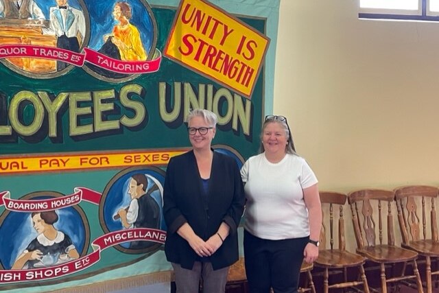Two smiling women stand in a hall in front of a historic union banner.