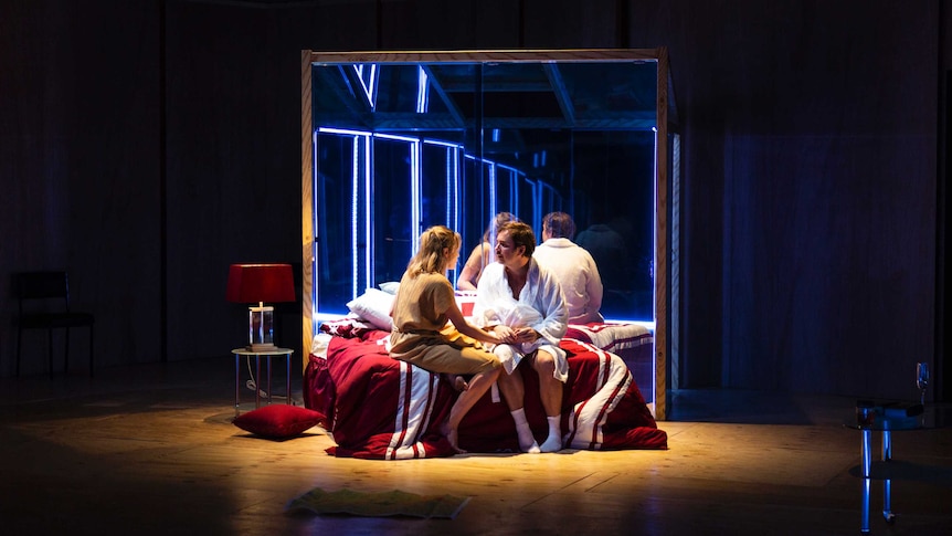 Anna Samson (left) and Toby Truslove (right) onstage sitting on a bed in front of a glasshouse.