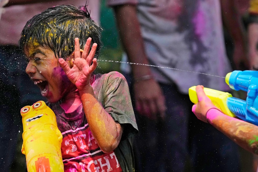 A young boy covered in coloured powder is being sprayed by a water gun as he looks away and laughs.
