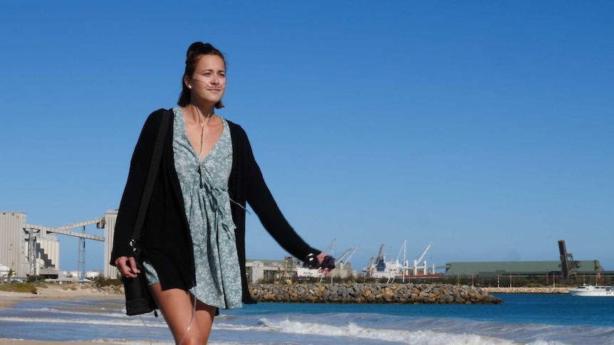 Cystic fibrosis ambassador Jackie Fraser walks along a beach in Geraldton, breathing oxygen delivered by a portable device.