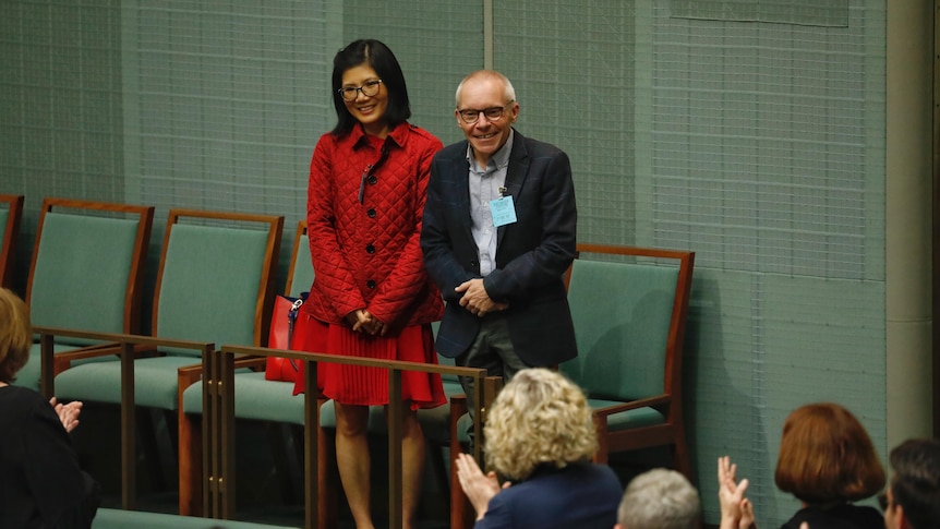 A woman of Vietnamese heritage in red and a Caucasian man in suit standing and smiling in parliament.