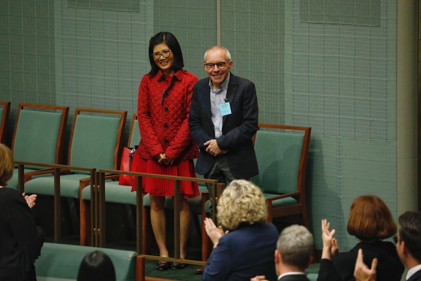 A woman of Vietnamese heritage in red and a Caucasian man in a suit standing and smiling in parliament.
