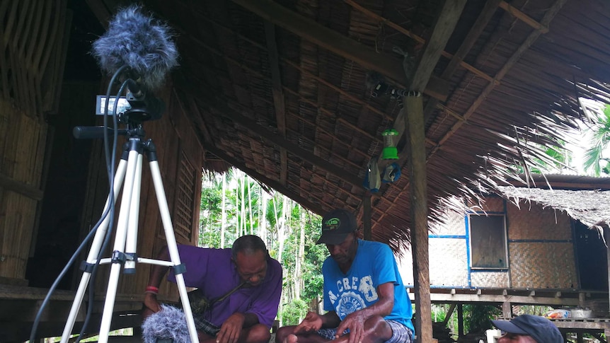 Men sit on verandah in wooden house look over pages in front of camera.