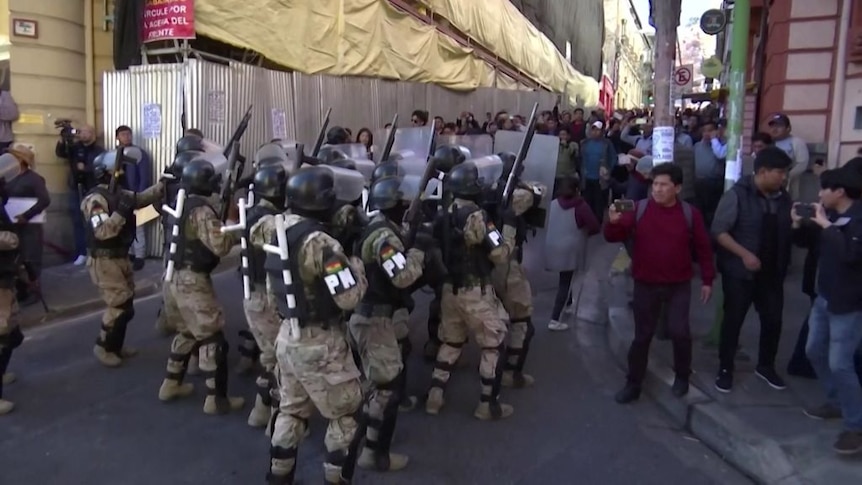 A group of armed troops in fatigues and riot gear are confronted by protesters on a city street.