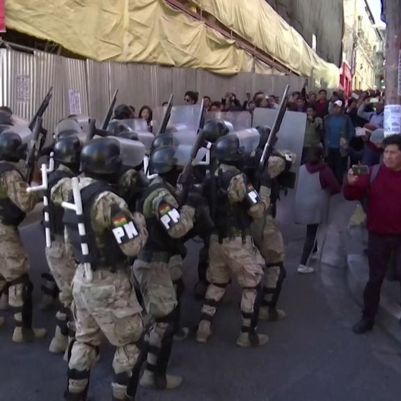 A group of armed troops in fatigues and riot gear are confronted by protesters on a city street.