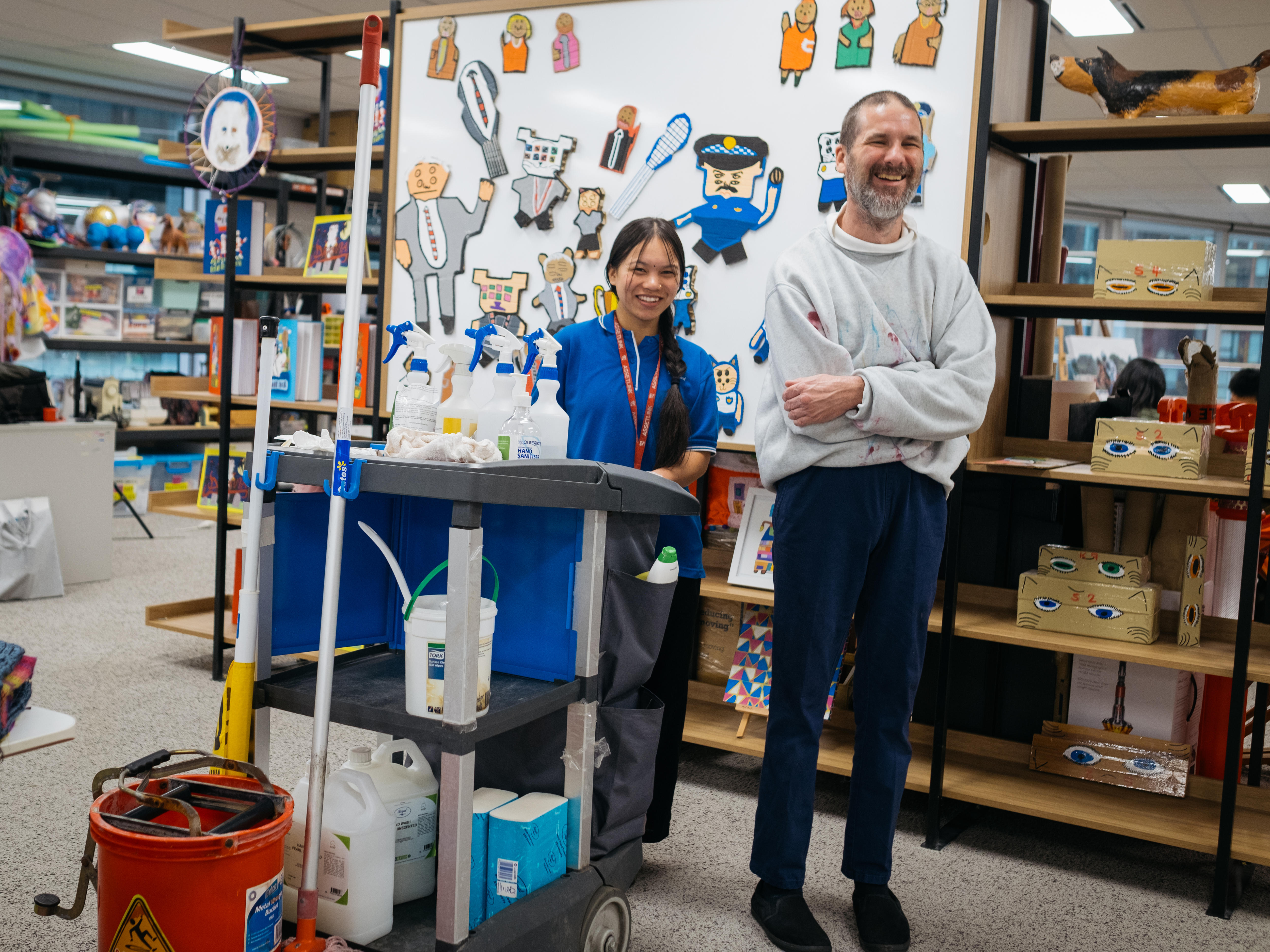 Cleaner Subita stands next to artist Thom in an art studio, they are both smiling. She is short with dark hair and a blue shirt