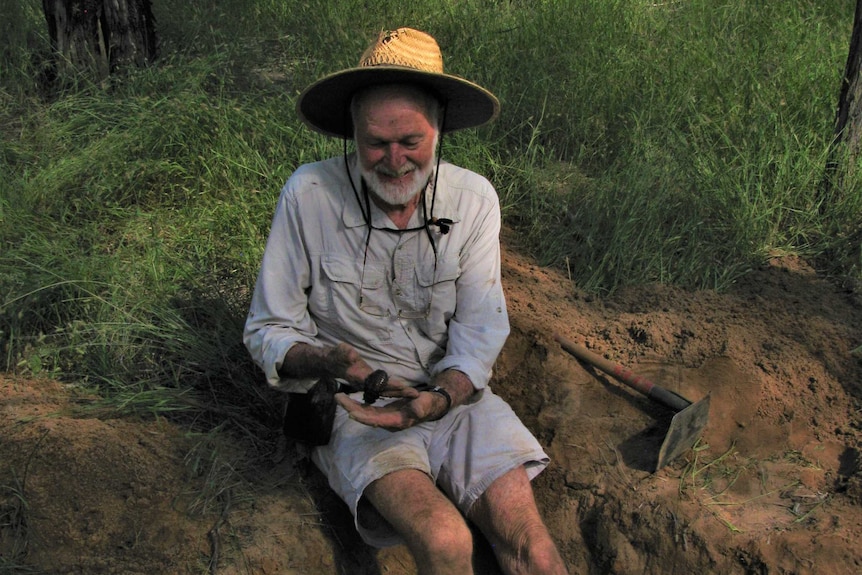 A man sits sit a dirt hole holding a giant cockroach.