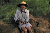 A man sits sit a dirt hole holding a giant cockroach.