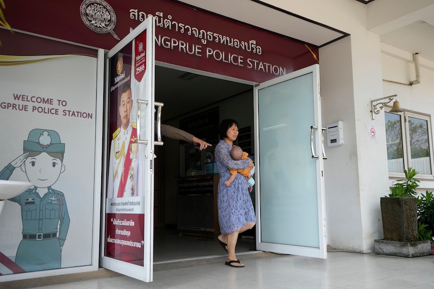 A woman in lavender dress walks out of police station holding baby in her arms. A hand is pointing out the door. 