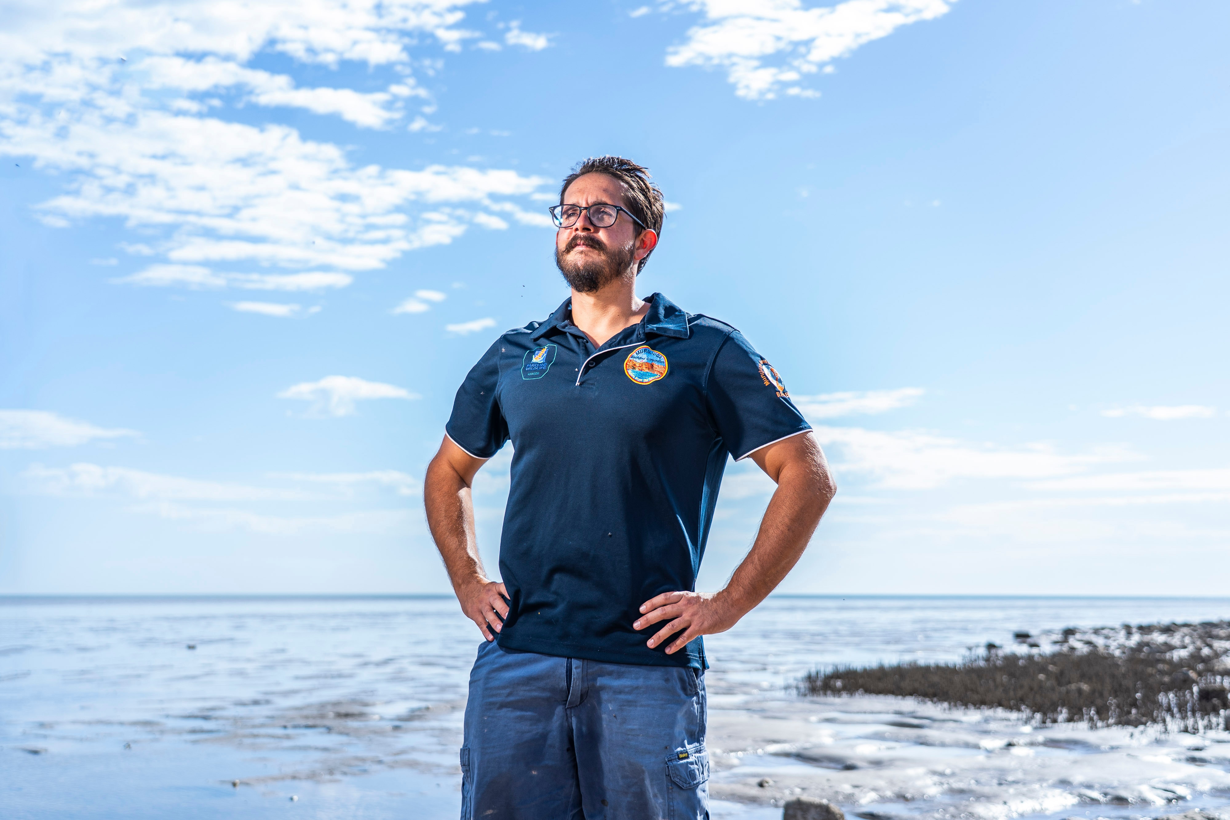 Um homem de óculos e blusa preta está com as mãos na cintura, com um céu claro e um oceano atrás dele.