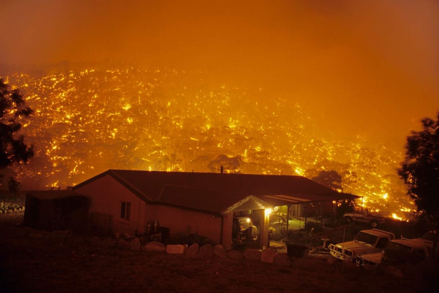 A homestead silhouetted by many fires behind it on the side of a hill