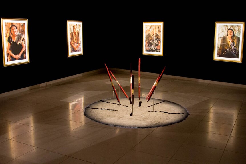 Four portraits of older women wearing animal furs hang in a black room. Spears arranged in circle in centre of room.