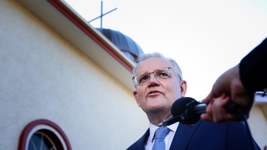Scott Morrison speaks into a microphone with the cross on top of a church behind him