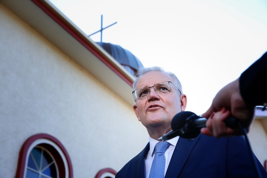 Scott Morrison speaks into a microphone with the cross on top of a church behind him