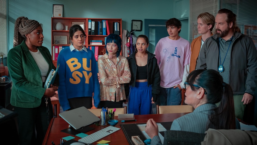 Five students and two teachers stand in front of a school principal at her desk.
