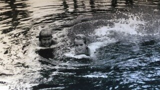 two young girls swim in a waterhole. They are holding onto paddle boards and smiling.