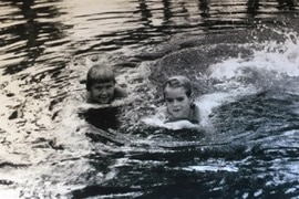 two young girls swim in a waterhole. They are holding onto paddle boards and smiling.