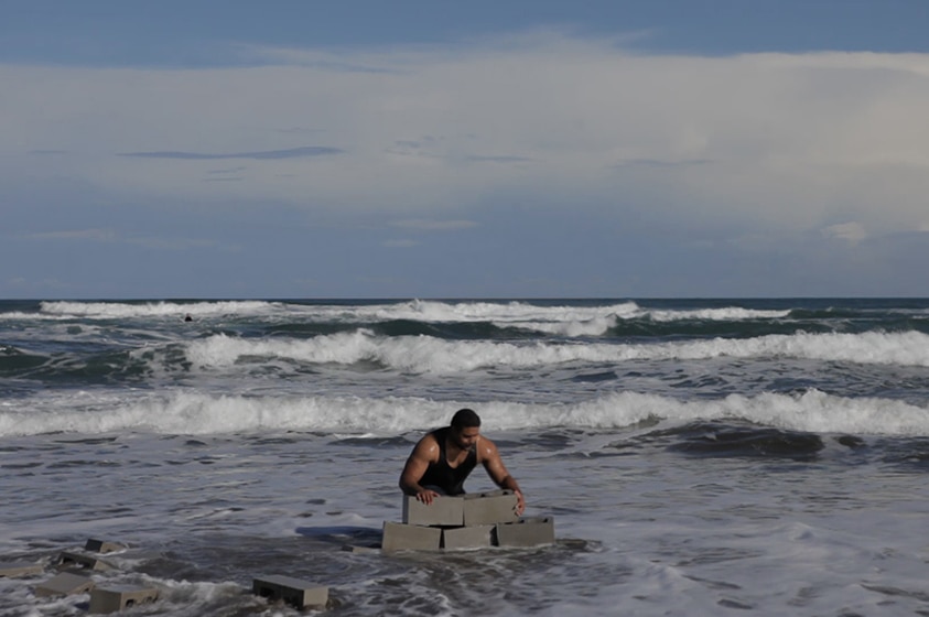 A man attempts to build a wall with cinderblocks on the beach as waves surround him on a bright day.