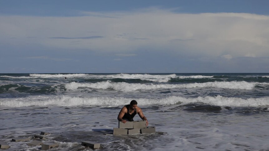 A man attempts to build a wall with cinderblocks on the beach as waves surround him on a bright day.