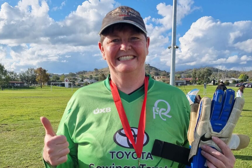 A short-haired beaming woman , cap, holds gloves, poses with her 2022 championship medal in her green goalkeeper uniform.