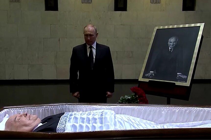 Man in suit stands beside a large black and white photo of a man, the same man who lies in a coffin in front of him.