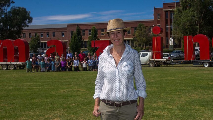 A woman standing on an oval with large red letters and people in the background in front of an old school building.