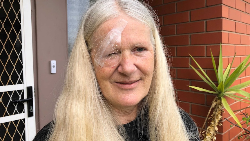 An older blonde woman standing at her front door wearing an eye patch.