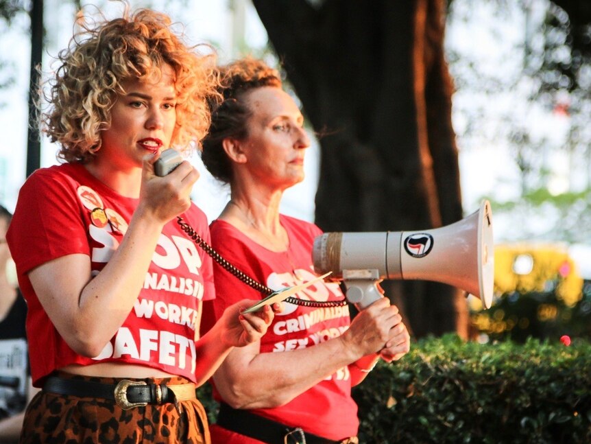 Two women in red shirts speak through a microphone at a rally.
