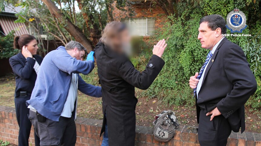 A man with his face blurred puts his hands in the air as a police officer pats him down.